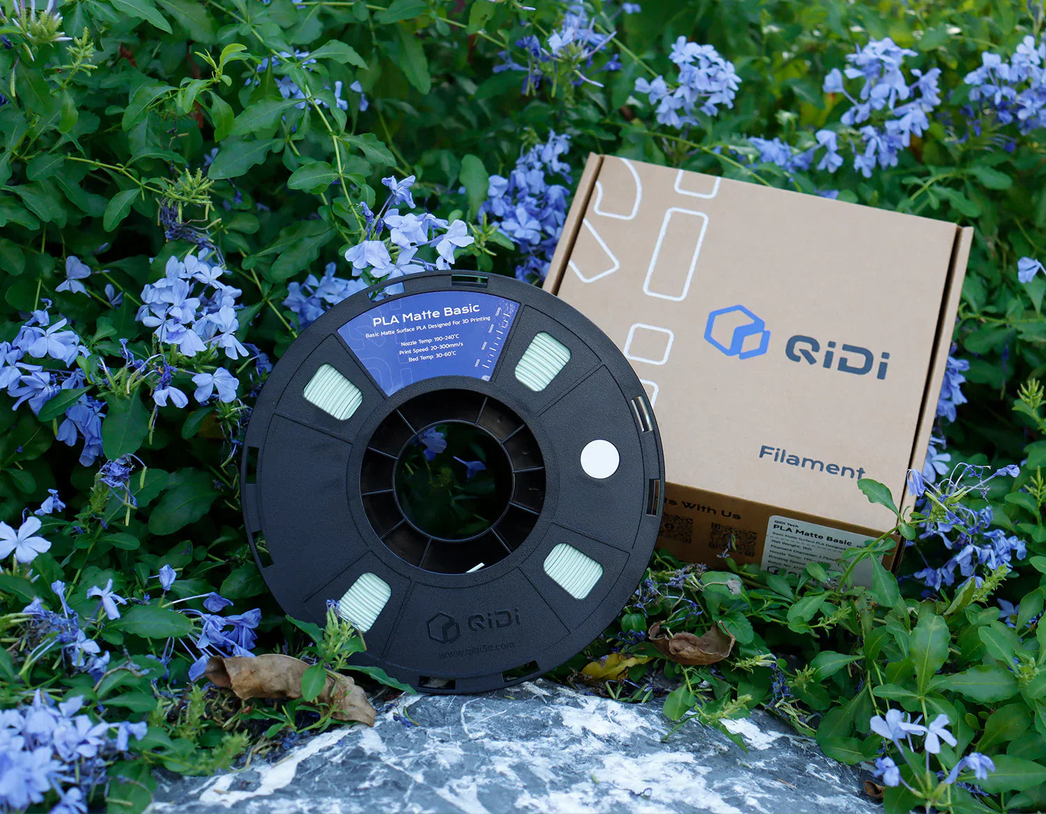 ブランドボックスに入った QIDI PLA 3D プリンター フィラメント スプール。屋外に紫色の花の間に展示されています。